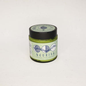 Medium size green jar of 'Nourish' Dry Skin Cream on a white background with a black lid. Three moons with a willow branch in dark blue on a light green label. 