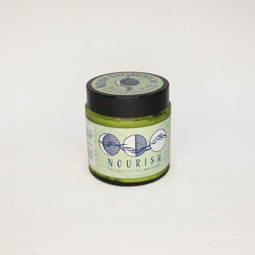 Medium size green jar of 'Nourish' Dry Skin Cream on a white background with a black lid. Three moons with a willow branch in dark blue on a light green label. 