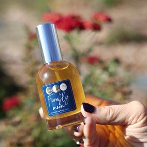 Hand holding a bottle labeled 'Firefly Moon' against a natural background with flowers.