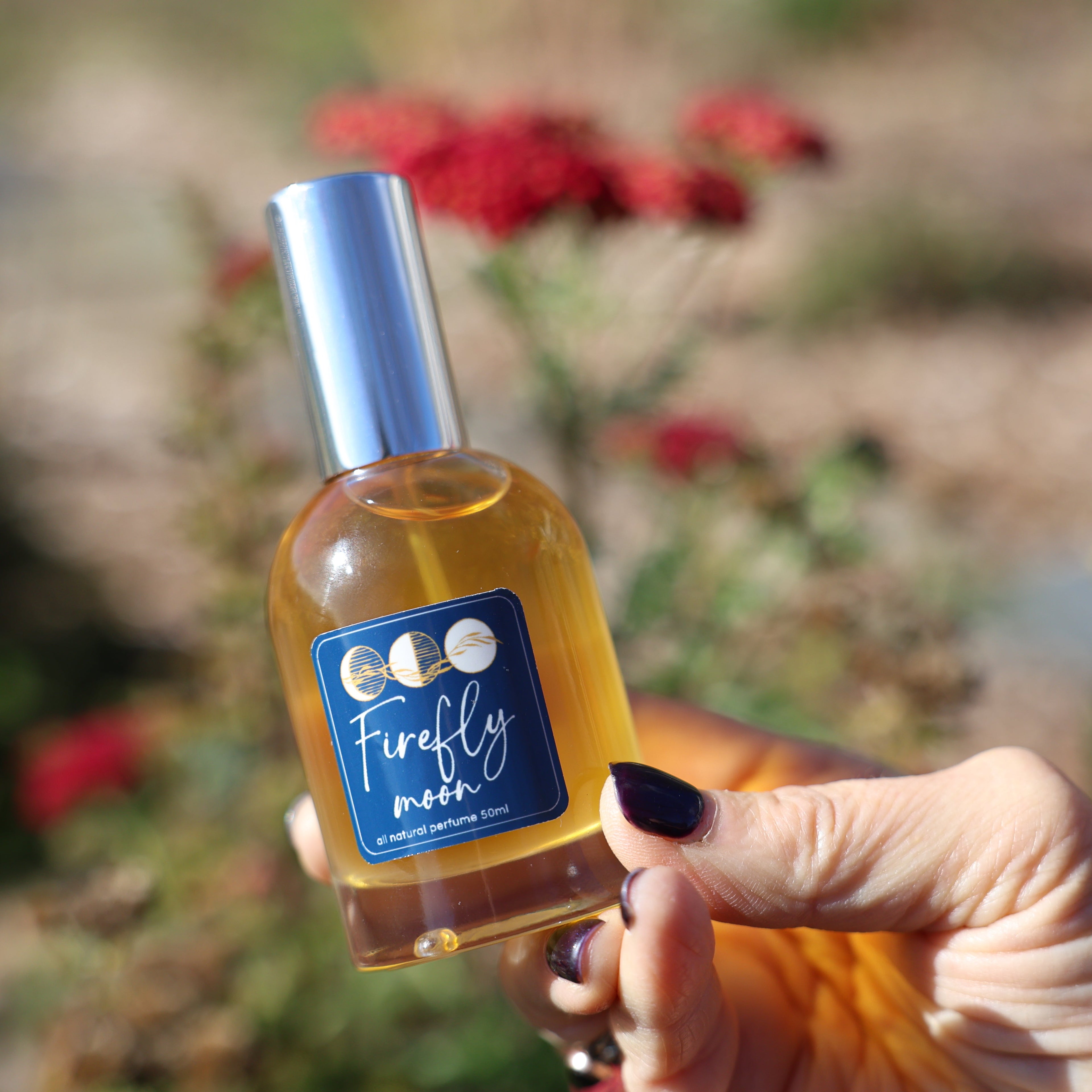 Hand holding a bottle labeled 'Firefly Moon' against a natural background with flowers.