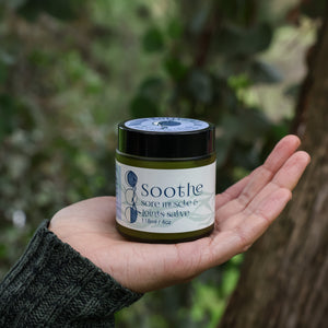 Hand holding a jar of 'Soothe Sore Muscle and Joints Salve' against a blurry background of deep green leaves and a brown tree trunk. 