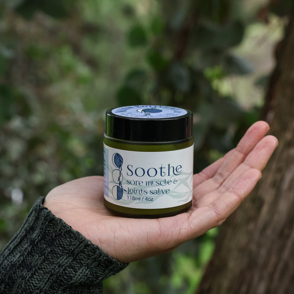Hand holding a jar of 'Soothe Sore Muscle and Joints Salve' against a blurry background of deep green leaves and a brown tree trunk. 